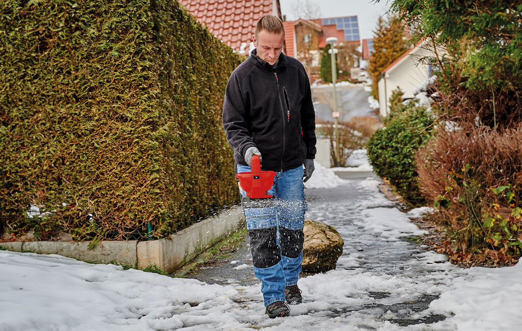Moški posipa poledenelo pot s pomočjo akumulatorskega trosilnega vozička in soli za posipanje.