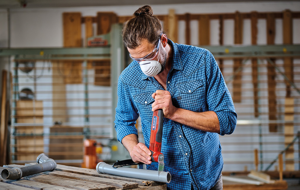 Moški reže plastično cev z Einhell multitool orodjem ter pri tem nosi zaščitna očala in masko.