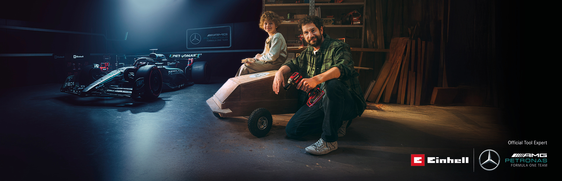 A father and child in a workshop with a homemade soapbox car. In the background, a Mercedes-AMG PETRONAS Formula One car and Einhell tools.