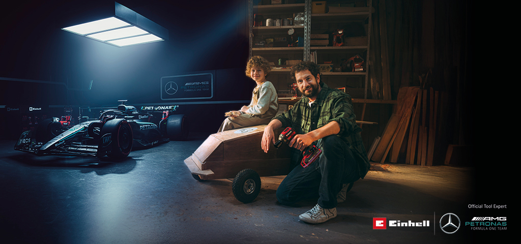A father and child in a workshop with a homemade soapbox car. In the background, a Mercedes-AMG PETRONAS Formula One car and Einhell tools.