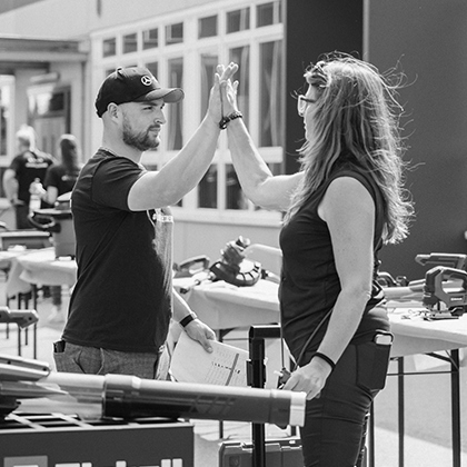 Muškarac i žena daju si high five na otvorenom Einhell događaju s izloženim alatima, prikazano u crno-bijeloj fotografiji.