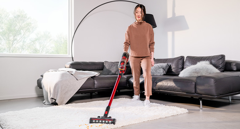 Frau saugt einen hellen Teppich im Wohnzimmer mit einem roten Akku-Staubsauger vor einem grauen Sofa.