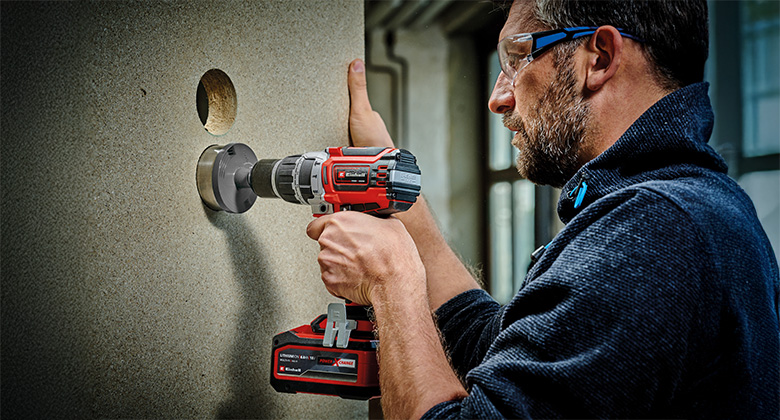 A man uses a cordless impact drill with a hole saw attachment to cut a large hole in a panel.