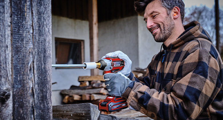 A man uses a cordless impact driver to drive a large screw into a wooden beam.